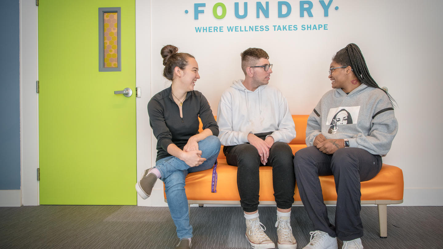 Three people sit on an orange couch in front of a sign that reads "Foundry"