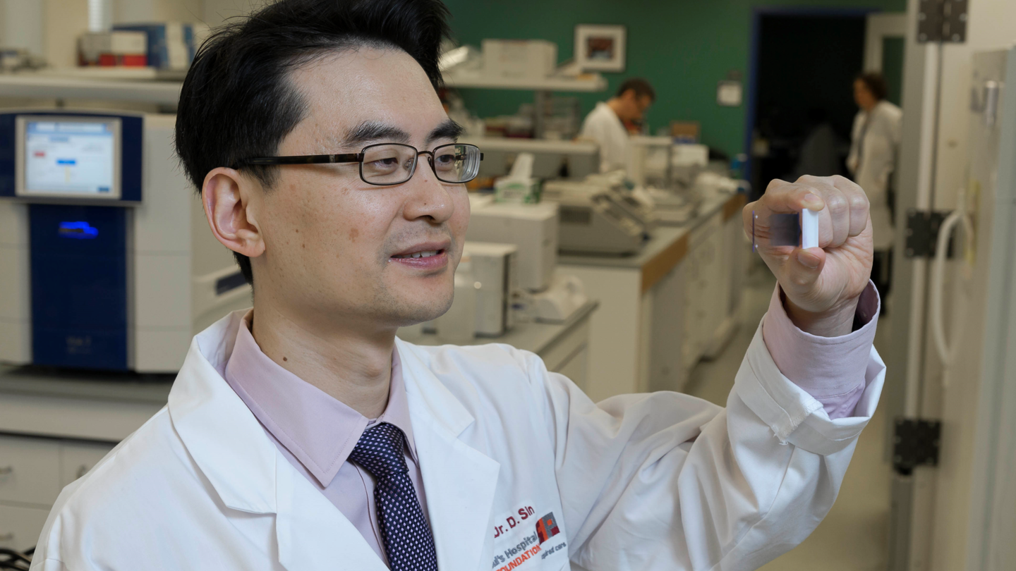 A man wearing a lab coat holds up a slide in the air while looking at it