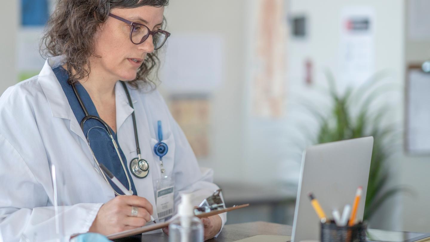 A woman in a lab coat writes on a clipboard while looking at a computer screen