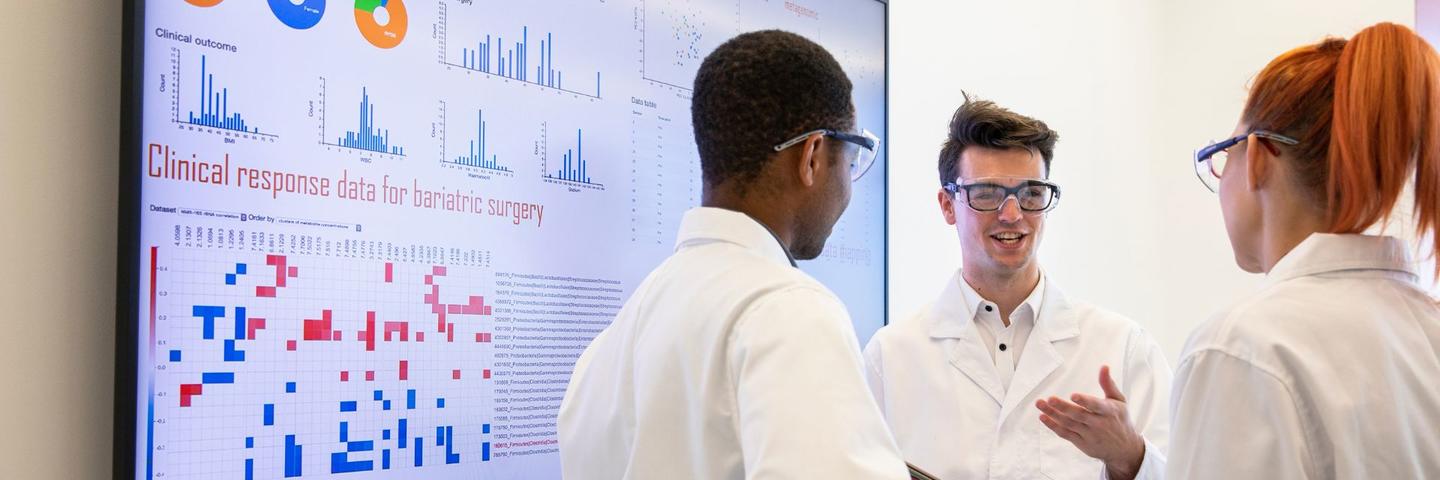 Three people in lab coats stand in front of whiteboard, chatting